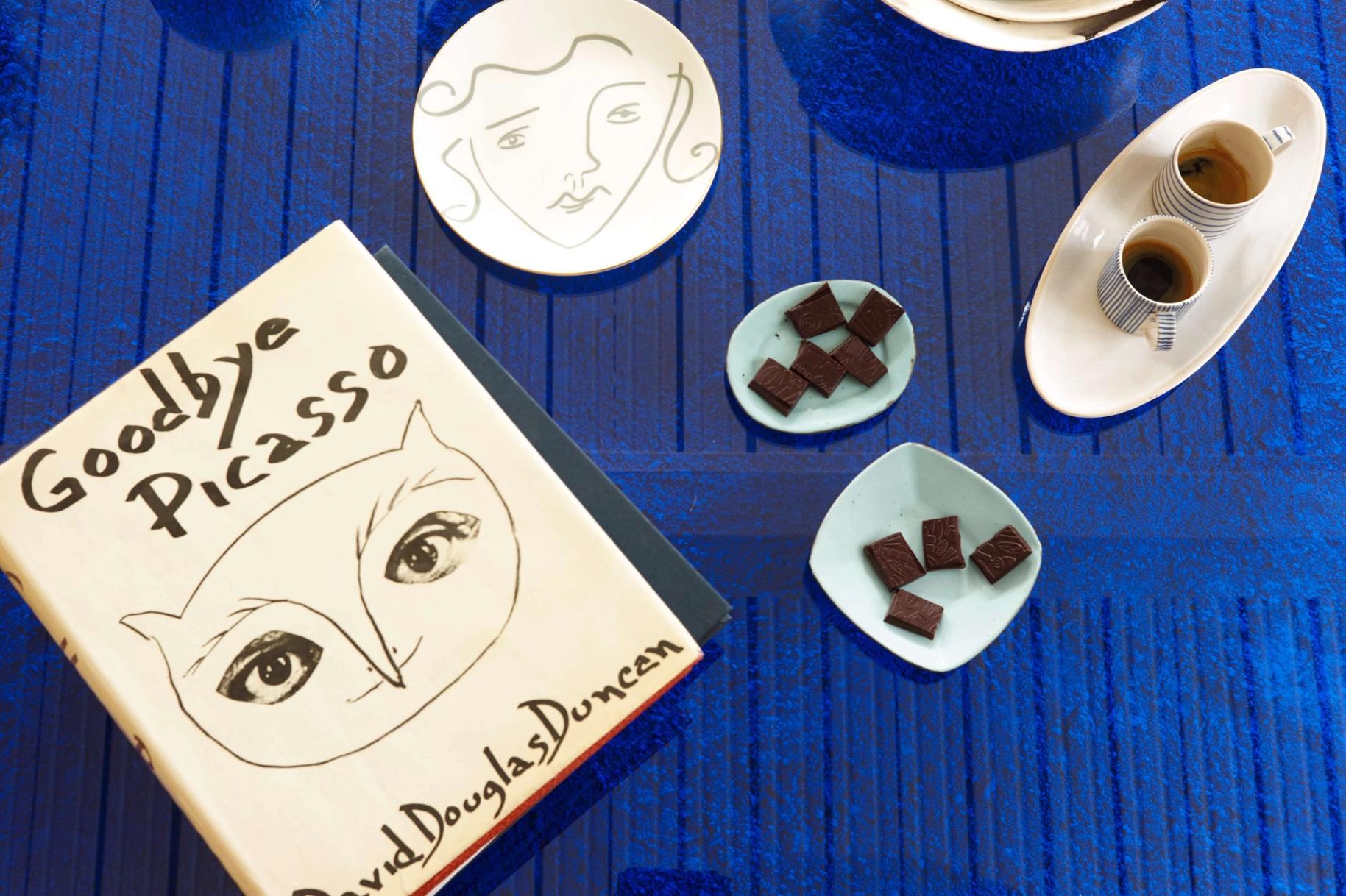 A book titled "Goodbye Picasso," a plate with a Picasso-esque face, two dishes of chocolates, and two espresso cups on a vibrant blue textured surface.