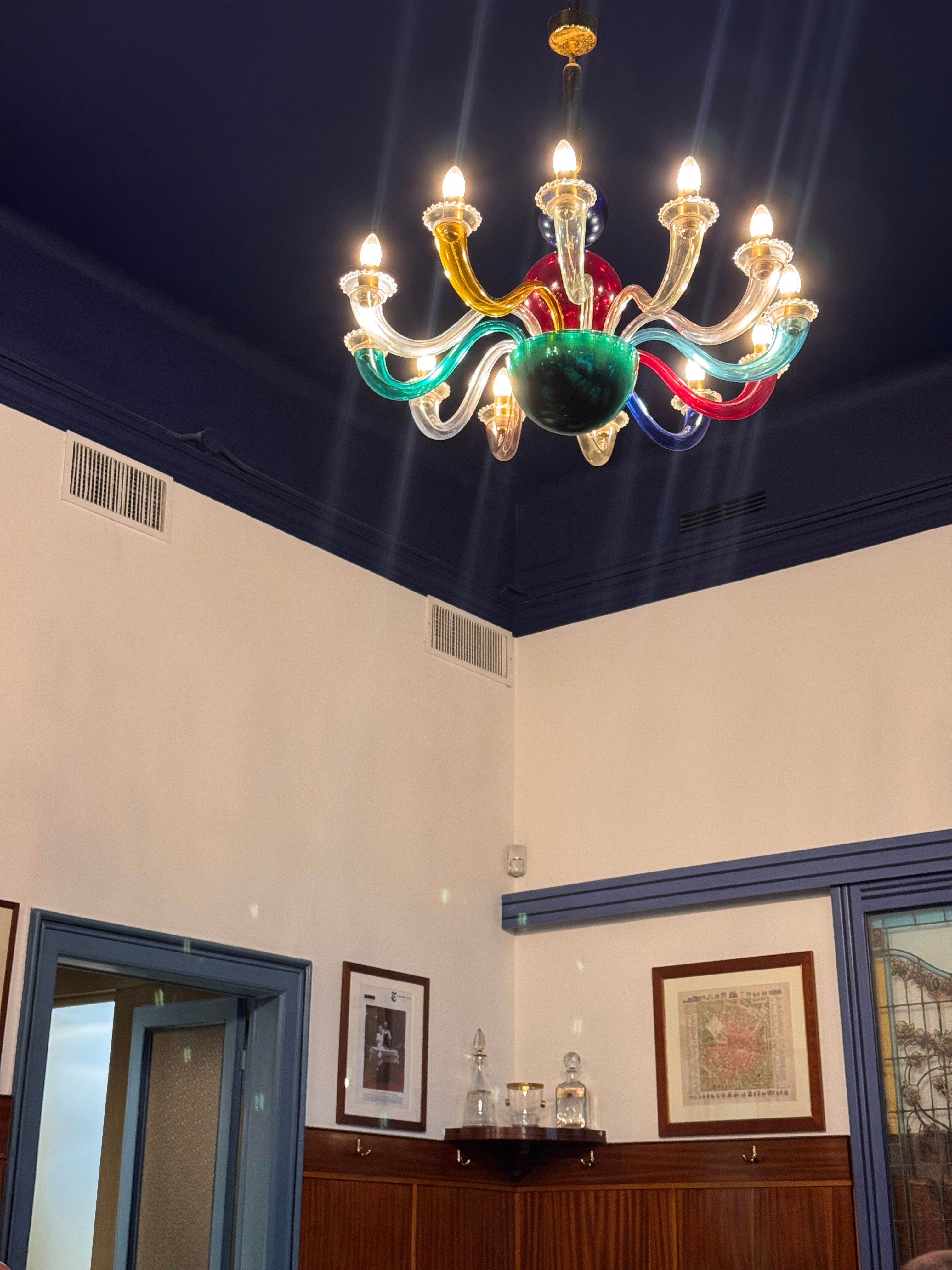 A bright, multicolored glass chandelier hangs from a dark blue ceiling above white walls and wood paneling.