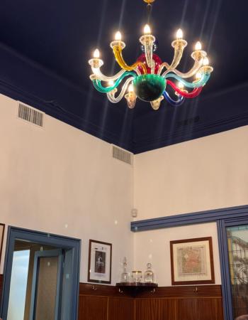 A bright, multicolored glass chandelier hangs from a dark blue ceiling above white walls and wood paneling.