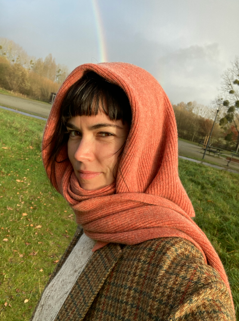 A woman wearing an orange hooded scarf and a tweed jacket stands outdoors with a rainbow in the sky.