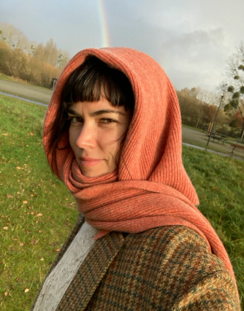 A woman wearing an orange hooded scarf and a tweed jacket stands outdoors with a rainbow in the sky.