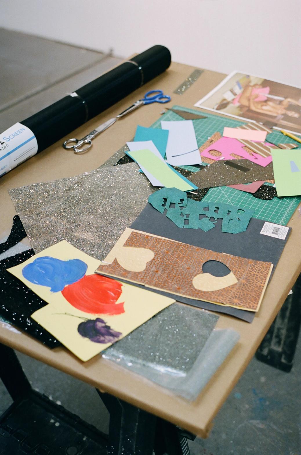 A craft table cluttered with art supplies including a rolled screen, scissors, colorful paper scraps, glitter paper, paint swatches, and a cutting mat.