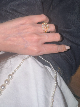 A hand wearing a gold heart-shaped diamond ring, with a pearl necklace draped over a white shirt and dark pants.