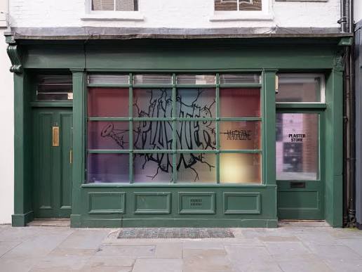 A dark green storefront with a large window displaying "PLASTER MAGAZINE" in black graffiti-style text over gradient colored panes, flanked by two green doors. The right door shows "PLASTER STORE" on a sign.