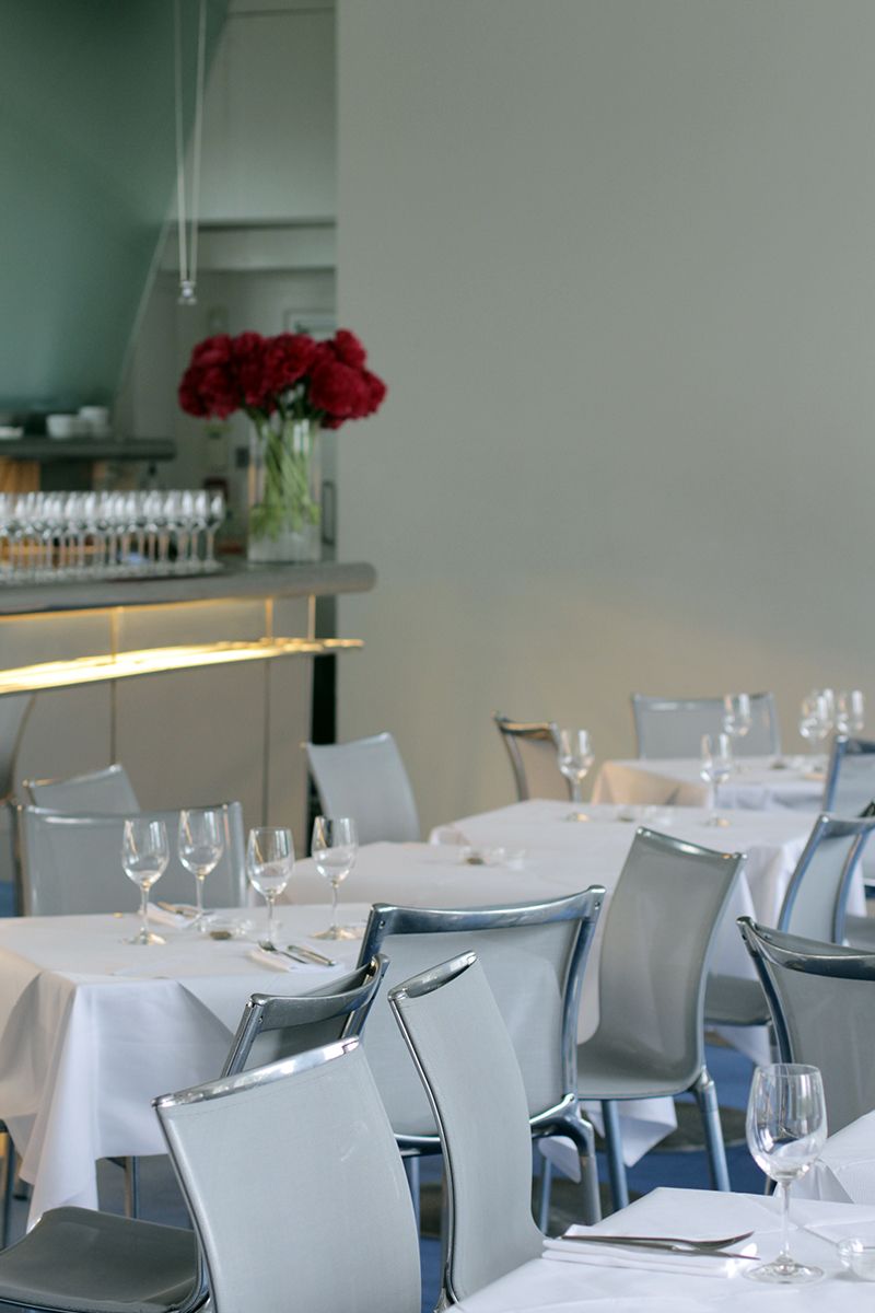 Modern restaurant dining room with white-clothed tables, grey chairs, and a bar with red flowers.