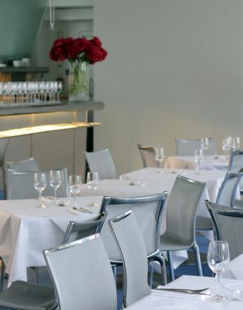 Modern restaurant dining room with white-clothed tables, grey chairs, and a bar with red flowers.