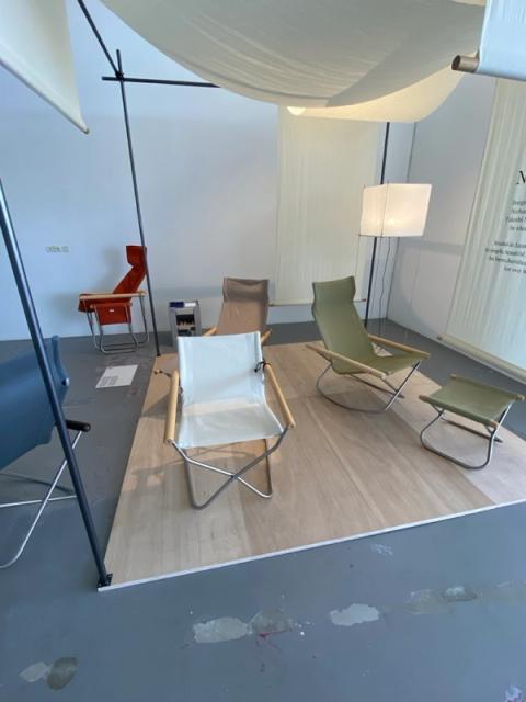 An indoor display of various minimalist folding chairs on a wooden platform, under a white fabric canopy.