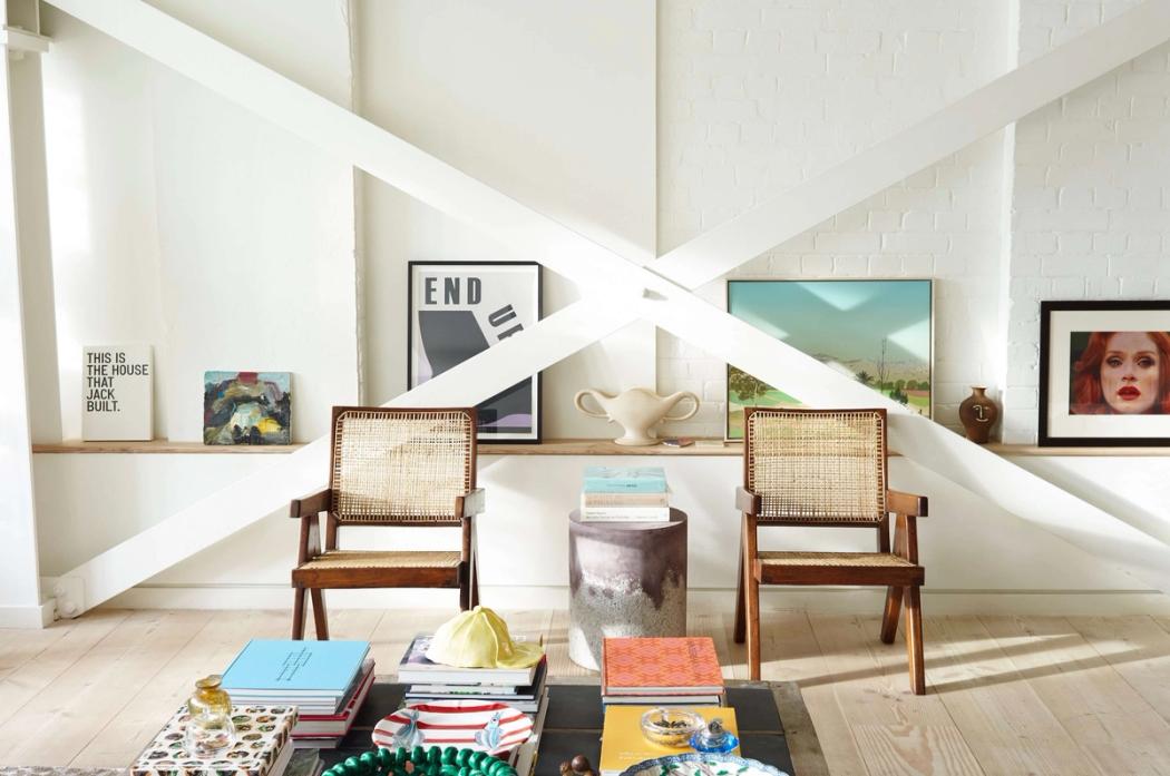 A bright room with two cane-backed chairs, a large white wall featuring intersecting diagonal beams, and shelves with art and books.