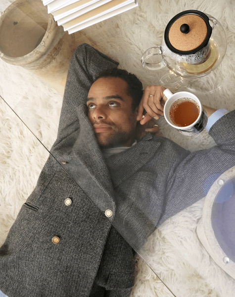 A man in a blazer lies on a white rug, looking up and holding a teacup, with a teapot and books nearby, seen through a glass surface.