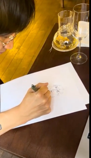 Overhead view of a person wearing glasses drawing with a pen on white paper, with drinks on the table.
