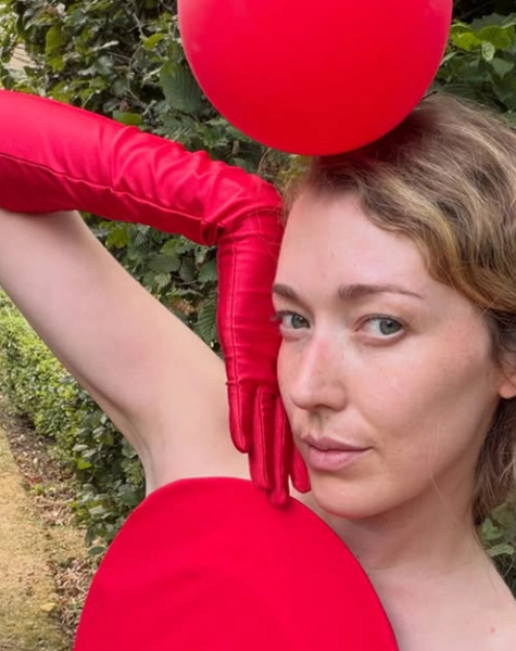 A bare-shouldered woman with wavy hair poses in a garden, with red gloves, a red balloon on her head, and a red circle covering her chest.