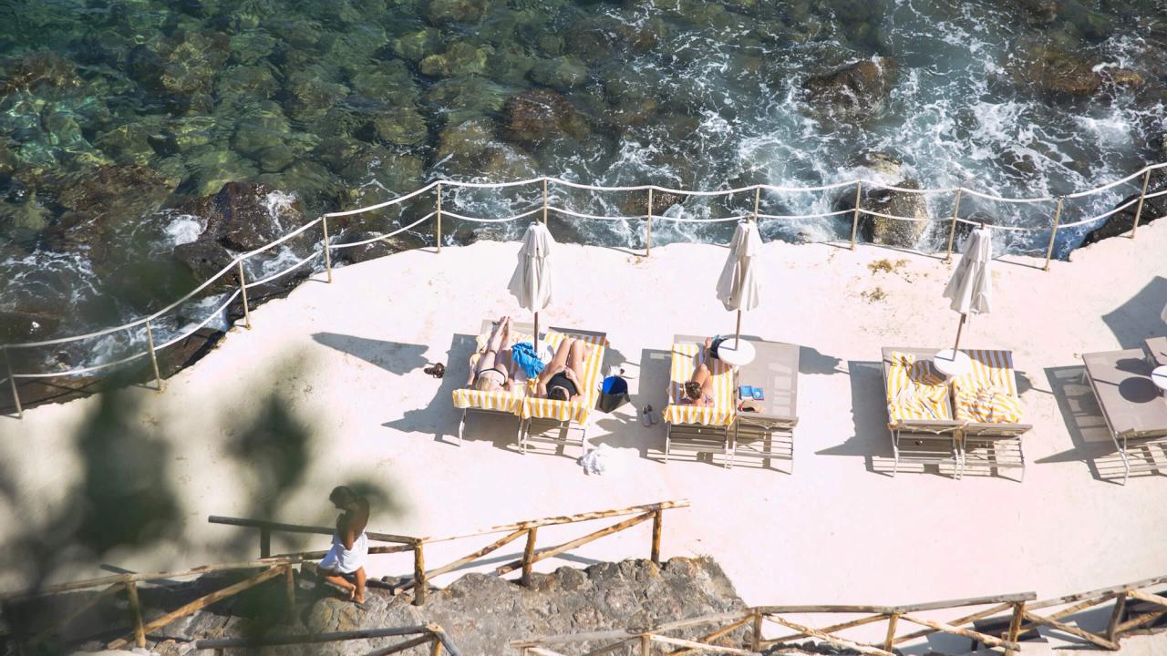 Overhead view of a sunny coastal leisure area with sunbathers on loungers by clear, rocky water.