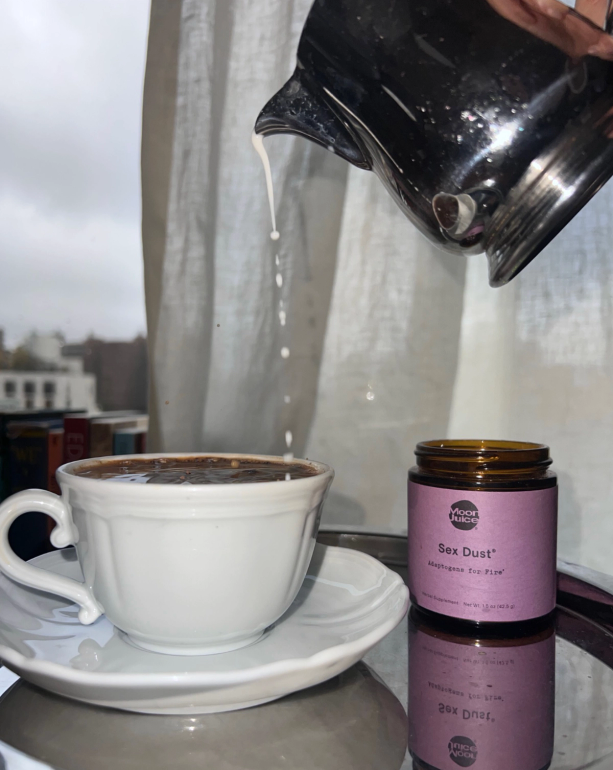 A hand pours milk into a white coffee cup next to a jar of Moon Juice Sex Dust, on a reflective surface by a window.
