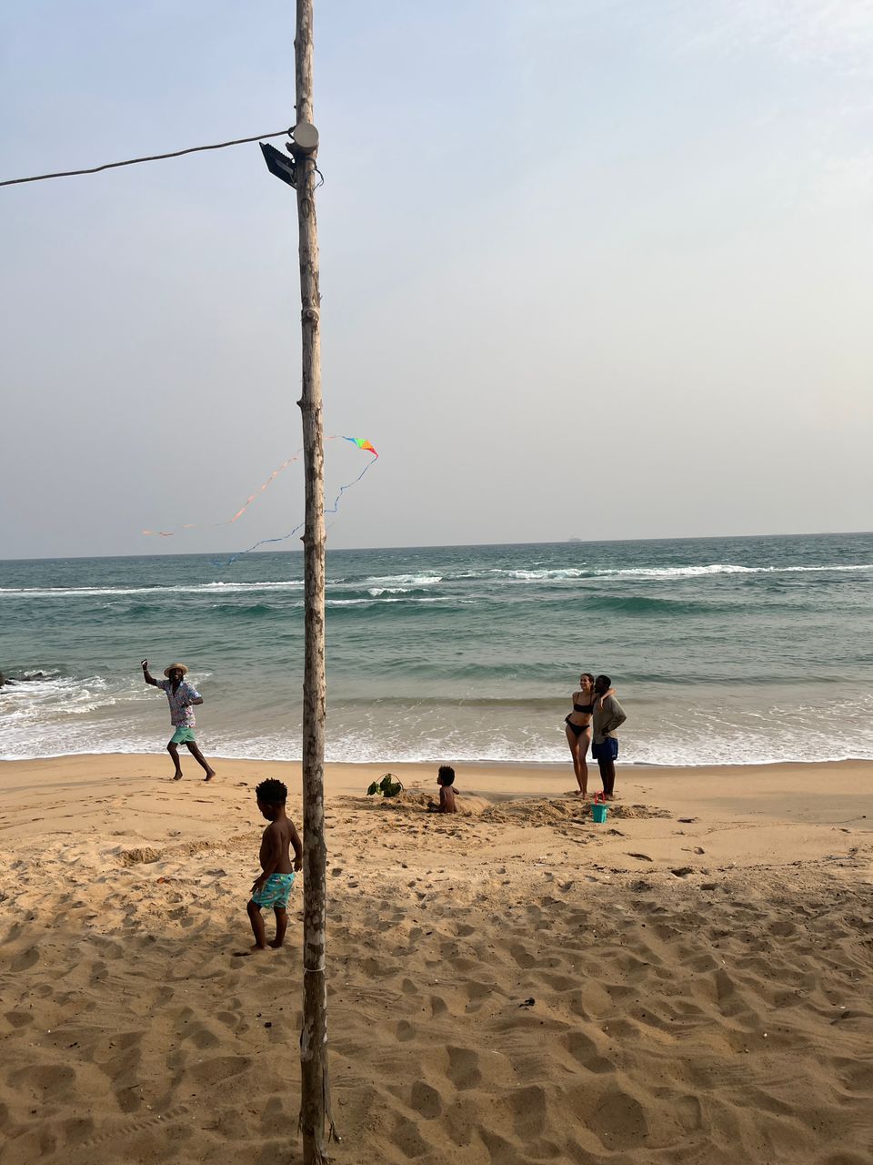 A beach scene featuring a tall wooden pole in the foreground, a man flying a kite, children playing in the sand, and a couple standing by the ocean.