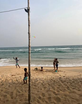 A beach scene featuring a tall wooden pole in the foreground, a man flying a kite, children playing in the sand, and a couple standing by the ocean.