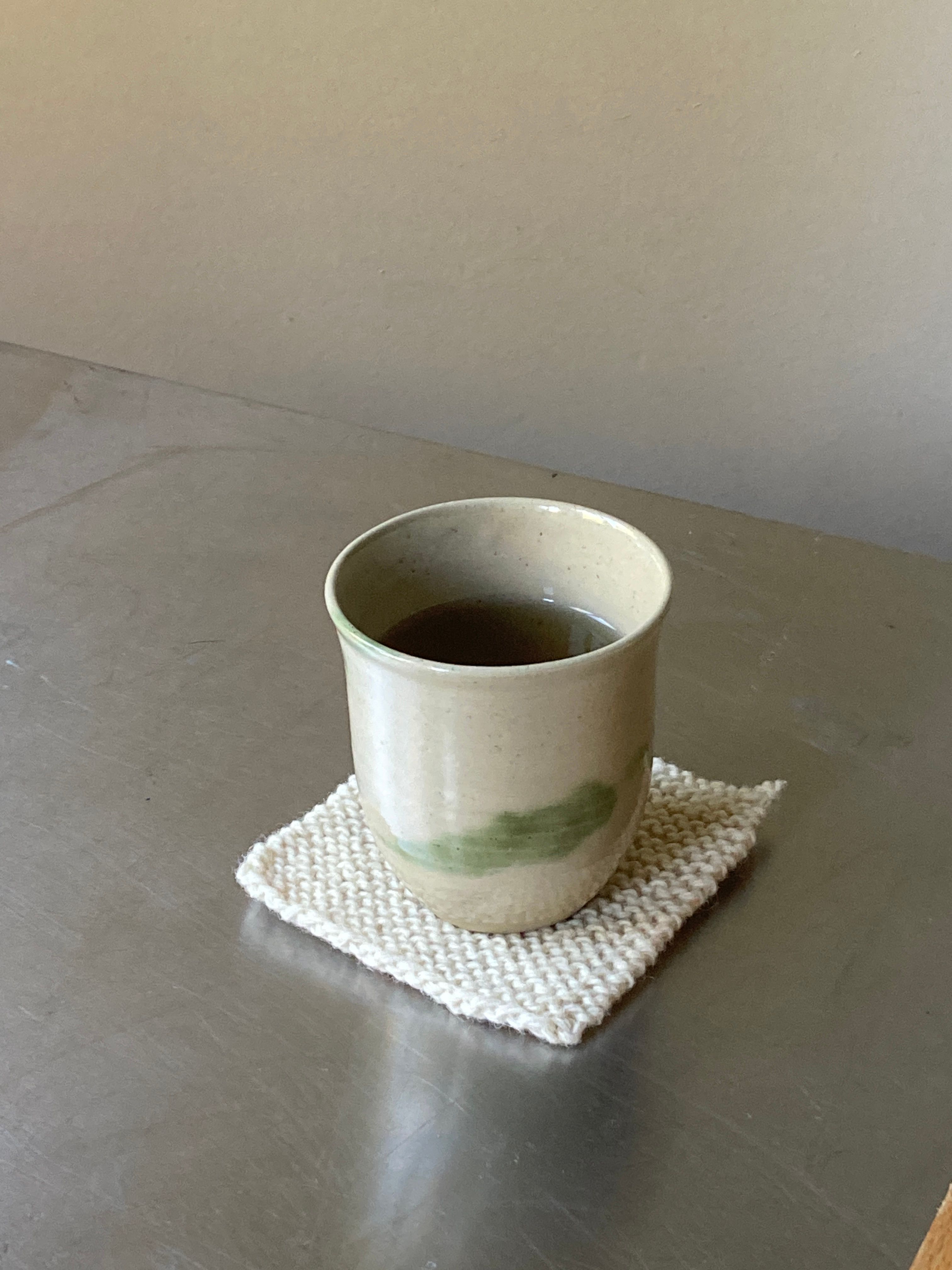 A light ceramic cup with a green brushstroke sits on a knitted coaster, filled with dark liquid, on a metal surface.