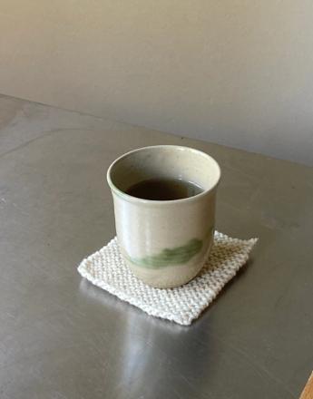 A light ceramic cup with a green brushstroke sits on a knitted coaster, filled with dark liquid, on a metal surface.