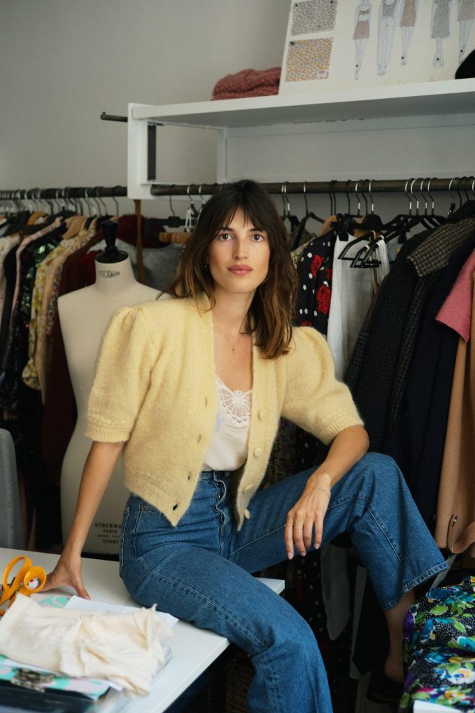 A woman with dark hair and bangs, wearing a yellow cropped cardigan and blue jeans, sits on a white table in a fashion studio with clothing racks.