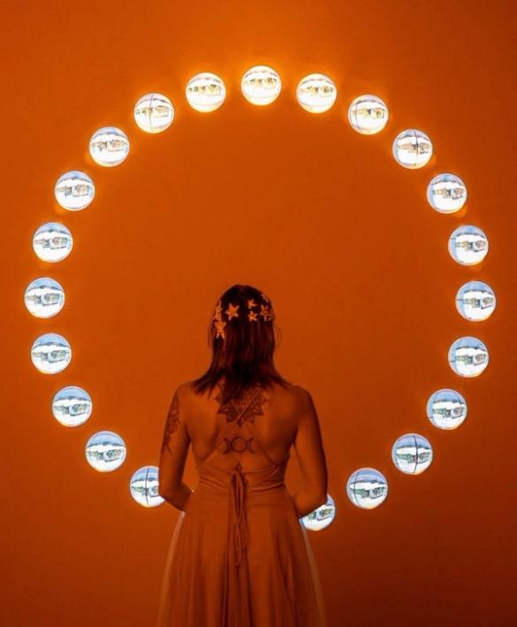 A woman with back tattoos and a star headpiece, seen from behind, stands before an orange wall featuring a large circle of glowing, reflective orbs.