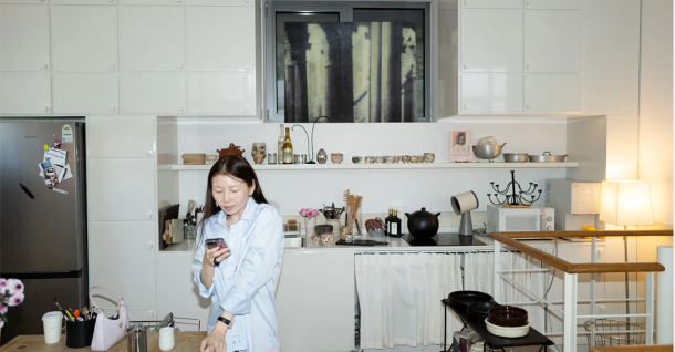 A woman in a light blue shirt stands in a white kitchen, looking at her phone.