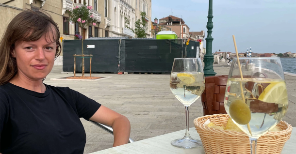 A woman sits at an outdoor table with two drinks, overlooking a canal.