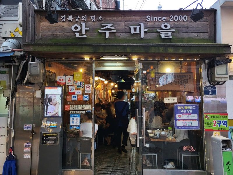 Front view of a bustling Korean restaurant called "Anju Maeul," with a rustic wooden sign and people dining inside.