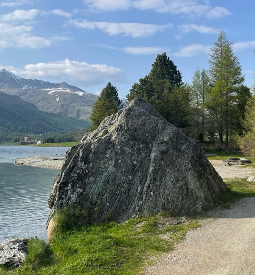 A large, lichen-covered rock stands by a lake, with snow-capped mountains, green trees, and a dirt path under a blue sky.
