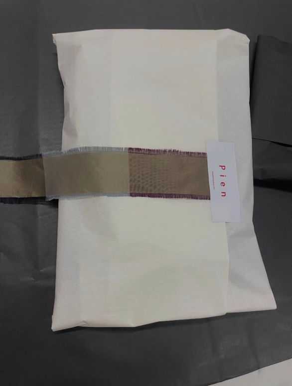 A package wrapped in white tissue paper, secured with a multi-toned brown and purple ribbon, and a "pien" brand tag.
