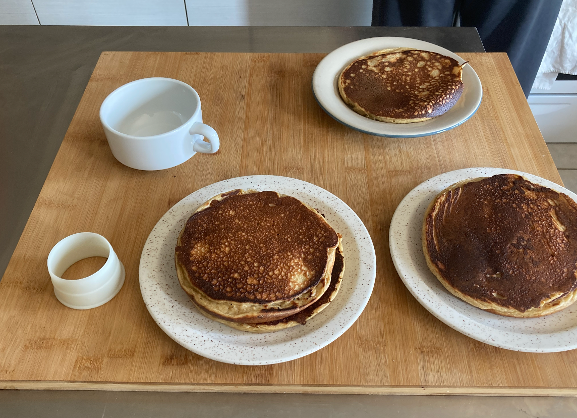 Three plates of pancakes, an empty white cup, and a white ring-shaped tool on a wooden cutting board.