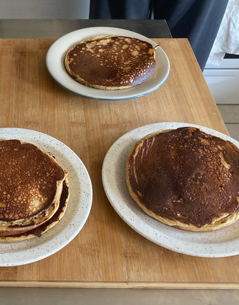 Three plates of pancakes, an empty white cup, and a white ring-shaped tool on a wooden cutting board.