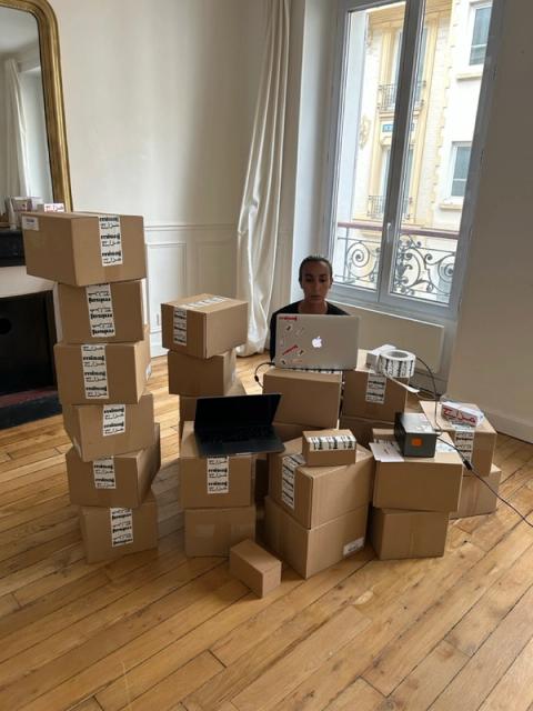 A woman sits working on a laptop, surrounded by many stacks of cardboard shipping boxes.