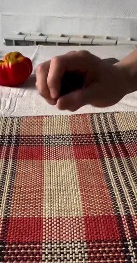 A hand grips a dark object over a red plaid placemat, with a red and yellow bell pepper to the left.