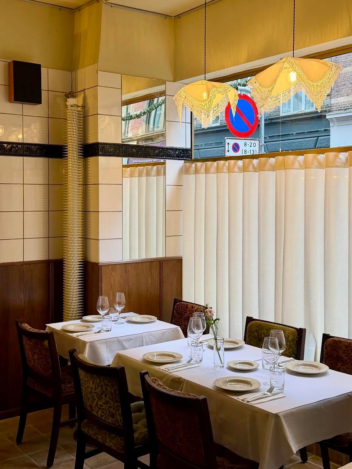 An empty restaurant interior with two white-clothed tables, tiled walls, wood paneling, and two lace pendant lights. A "no stopping" sign is visible outside a window.