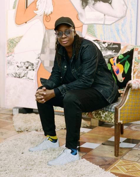 A Black person with dreadlocks, wearing a cap and denim jacket, sits on a colorful armchair in an eclectic room with artwork and patterned tiles.