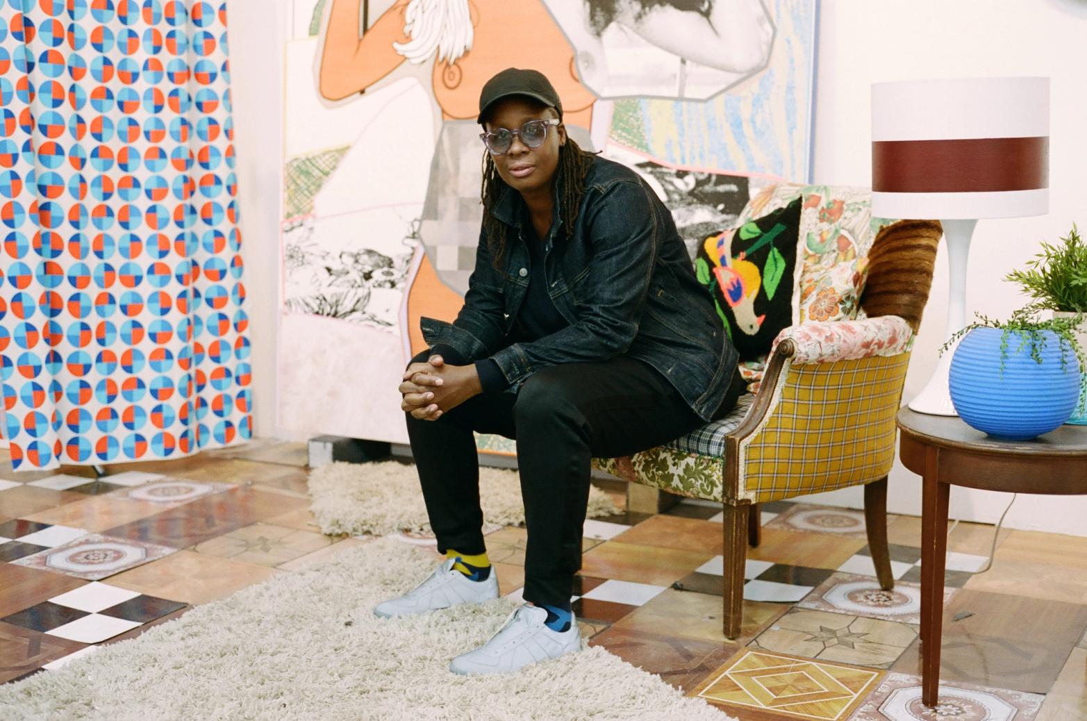 A person with dreadlocks wearing a denim jacket and cap sits on a patterned armchair in a colorful room with a large painting.