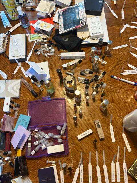 Numerous perfume bottles, sample vials, and labeled testing strips spread across a wooden table.