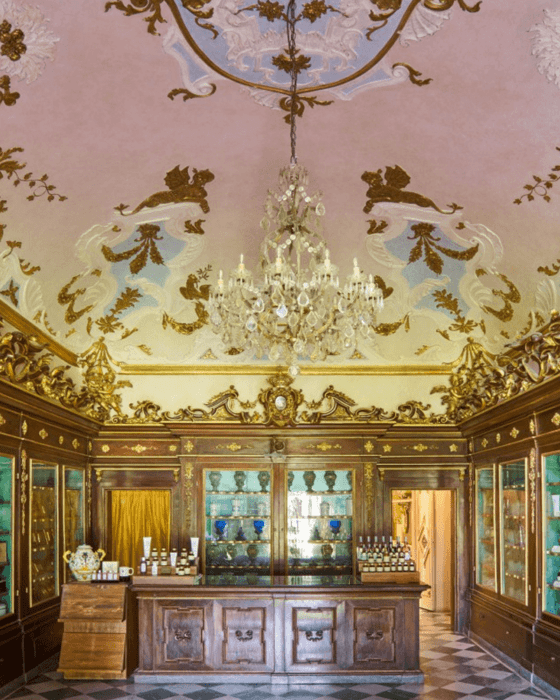 Elaborate historical apothecary interior with dark wood display cases, a counter, a large crystal chandelier, and an ornate pink and gold ceiling.