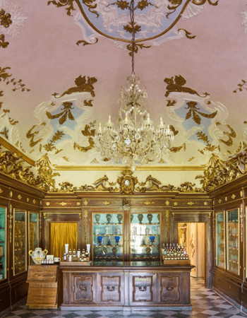 Elaborate historical apothecary interior with dark wood display cases, a counter, a large crystal chandelier, and an ornate pink and gold ceiling.