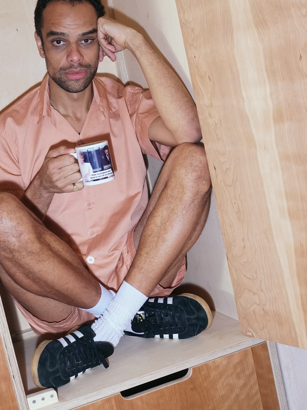 A man in a peach outfit and sneakers sits in a wooden cubby, holding a meme mug.
