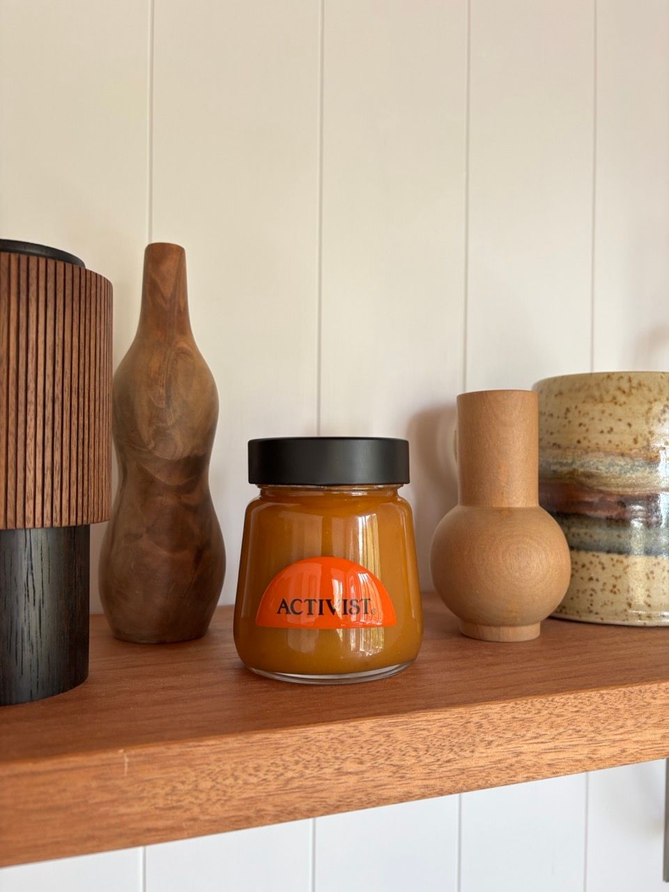 An "ACTIVIST" jar with orange contents sits on a wooden shelf surrounded by decorative wooden and ceramic objects.