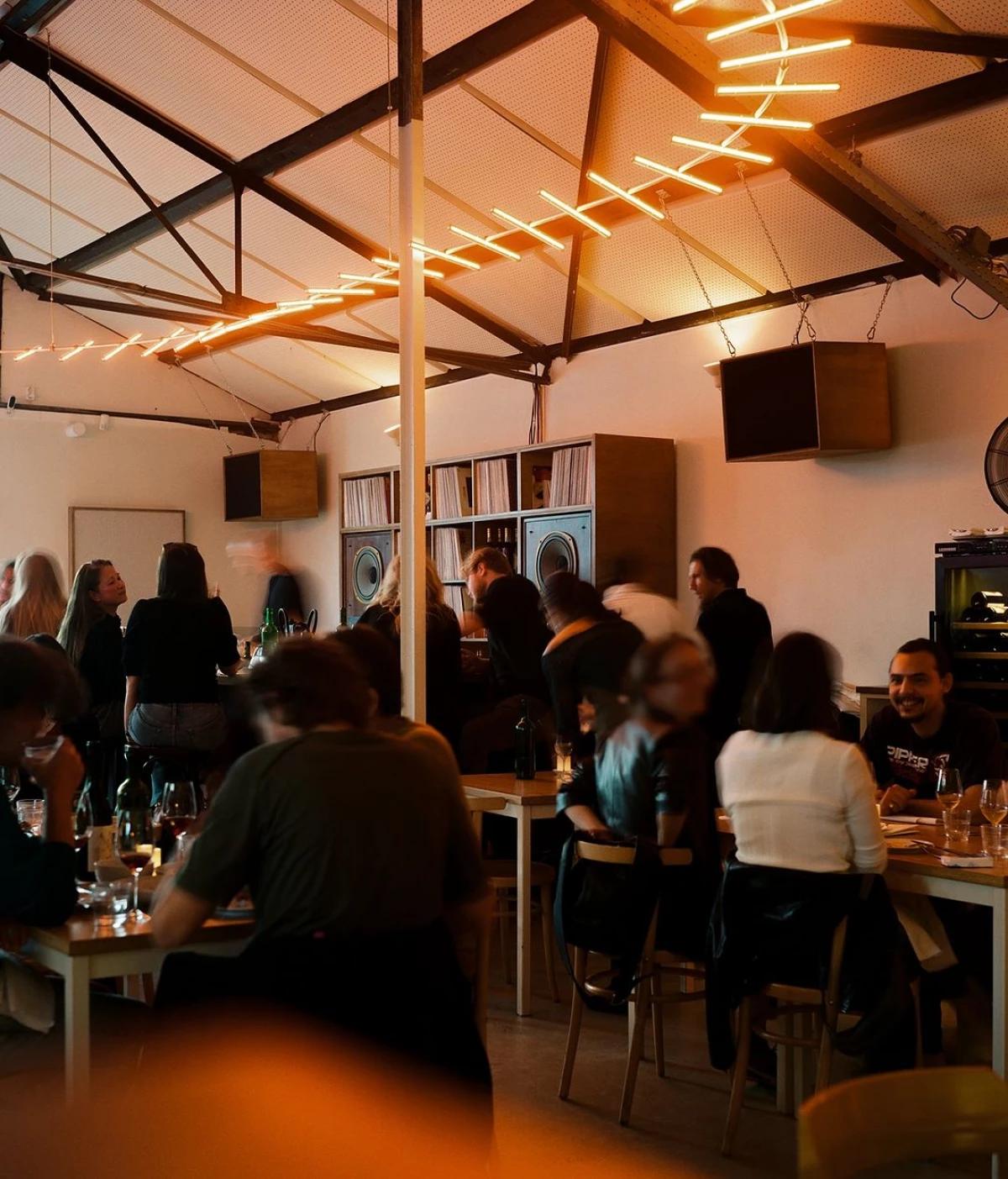 Restaurant interior with people dining under a distinctive orange linear light fixture, exposed beams, and a record collection.