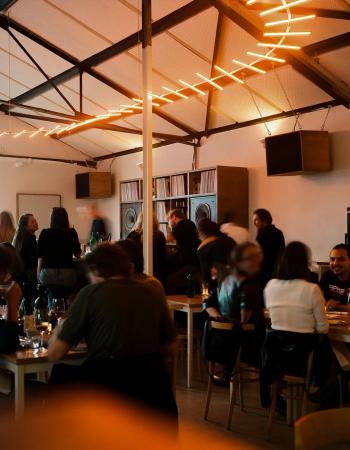 Restaurant interior with people dining under a distinctive orange linear light fixture, exposed beams, and a record collection.