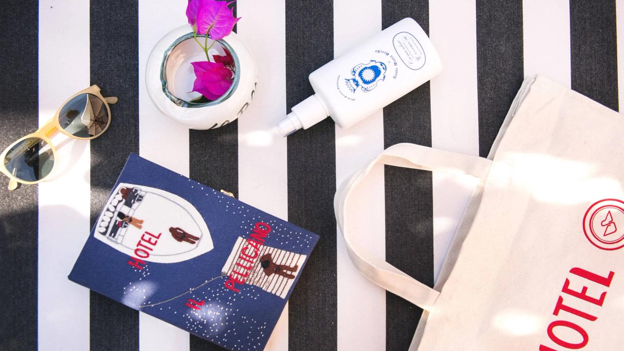 Flat lay of summer essentials: sunglasses, sunscreen, a vase with pink flowers, and Hotel Pelicano branded clutch and tote bags on a black and white striped surface.