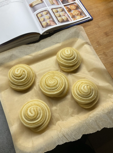 Six unbaked spiraled buns on parchment paper with a cookbook in the background.