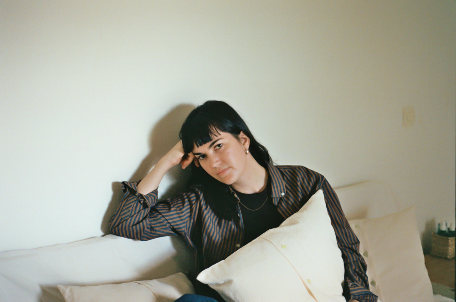A woman with dark hair and bangs sits on a couch, head propped on her hand, holding a pillow, wearing a striped shirt.