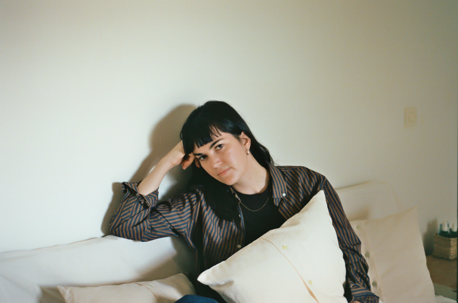 A woman with dark hair and bangs sits on a couch, head propped on her hand, holding a pillow, wearing a striped shirt.