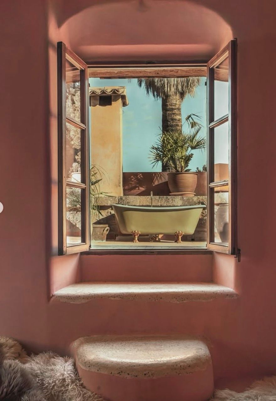 An open window in a pink room frames an outdoor terrace with a light green clawfoot tub and palm trees.