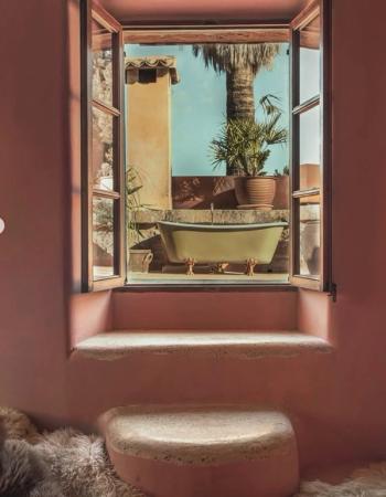 An open window in a pink room frames an outdoor terrace with a light green clawfoot tub and palm trees.