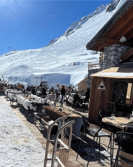A lively outdoor restaurant terrace on a snowy mountain, with many people dining at tables with furry white chair covers, next to a stone and wood chalet, all under a clear blue sky.