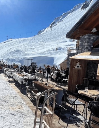 A lively outdoor restaurant terrace on a snowy mountain, with many people dining at tables with furry white chair covers, next to a stone and wood chalet, all under a clear blue sky.